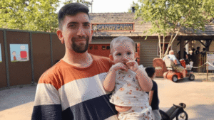 Father holding baby at theme park with Candy Cabin attraction in background.