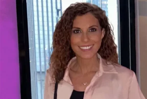 Smiling professional woman with curly hair in pink blazer standing in modern office with city views.