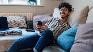 Smiling man with glasses relaxing on couch, holding smartphone, with laptop on nearby coffee table